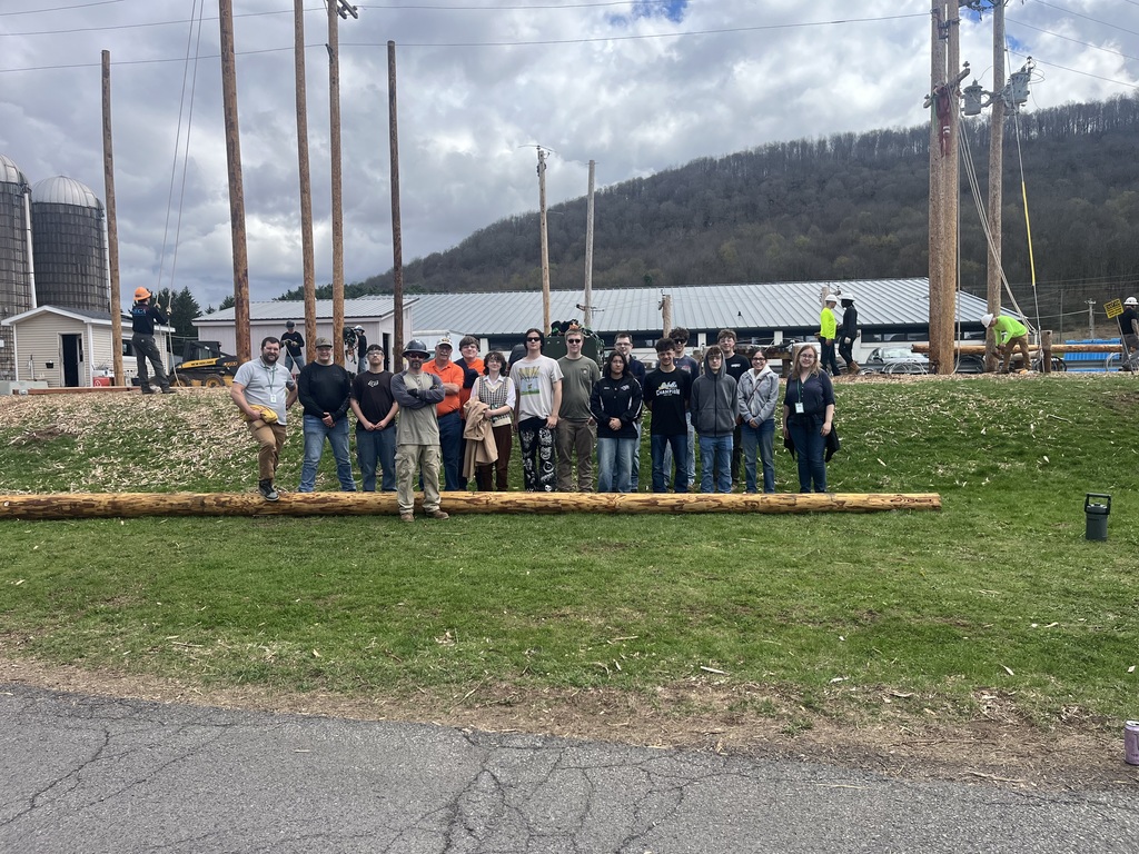 Mechatroni s students and teachers standing behind a large electric pole with college students and instructors in the background with standing electric poles.