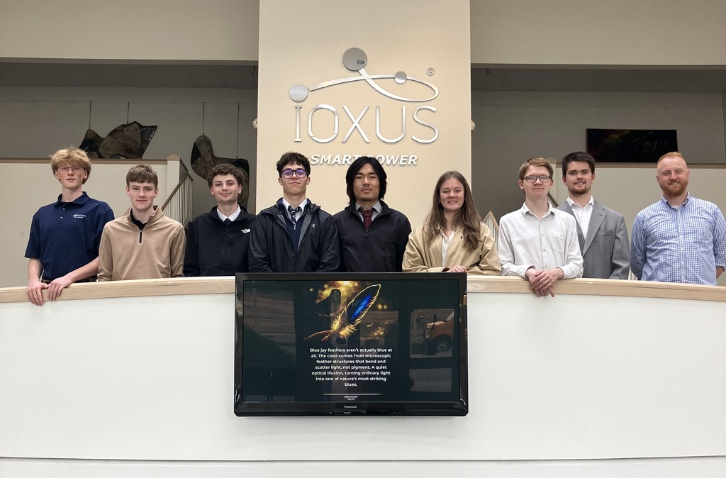Nine students standing in front in a building with the Ioxus sign behind them.