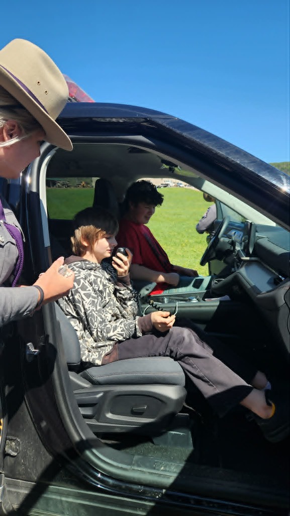 Two students sitting inside the front of a NYS Trooper vehicle with the tropper standing outside of it