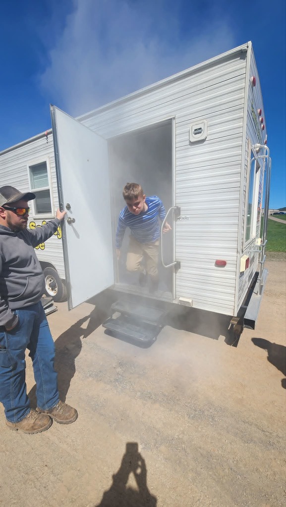 A firefighter is holding the door open to the smoke trailer while an elementary student is leaving it