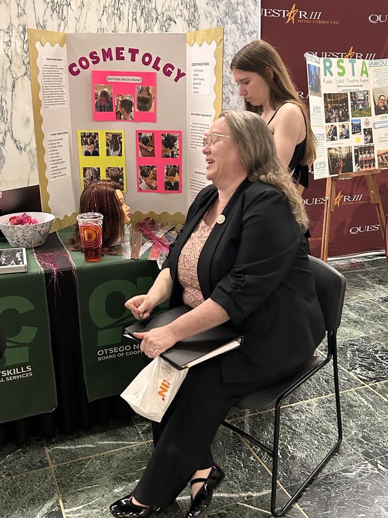 A visitor sitting in a chair near the cosmetology table while a student works on putting tinsel in their hair