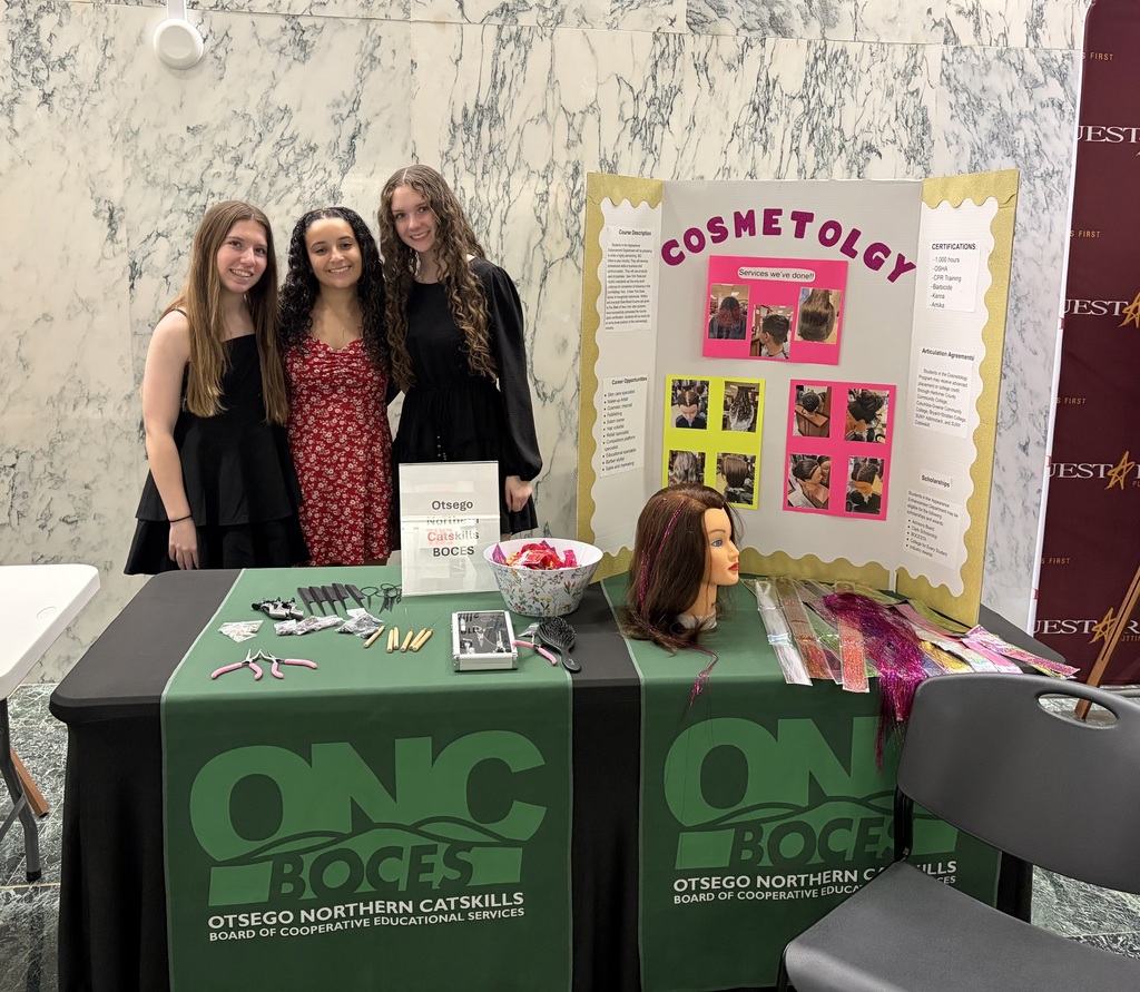Three cosmetology students posing behind a table with hair styling tools, mannequin head, and their presentation board
