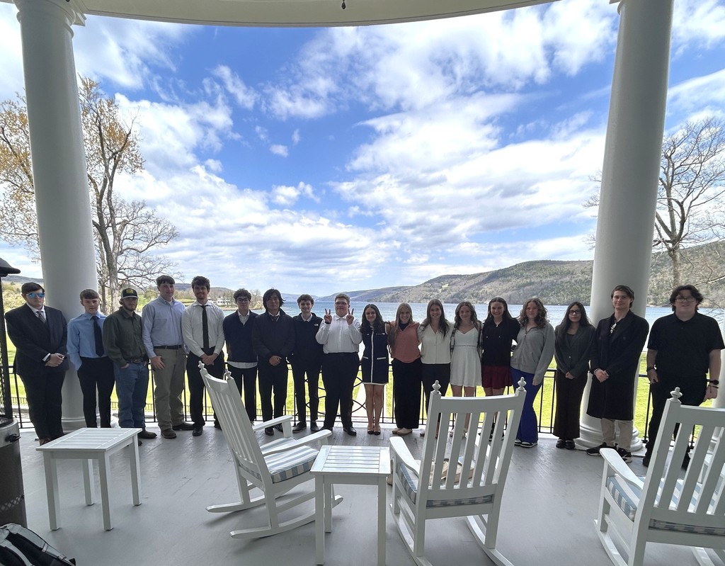 18 students standing together on a porch with white rocking chairs with a lake and mountains behind them