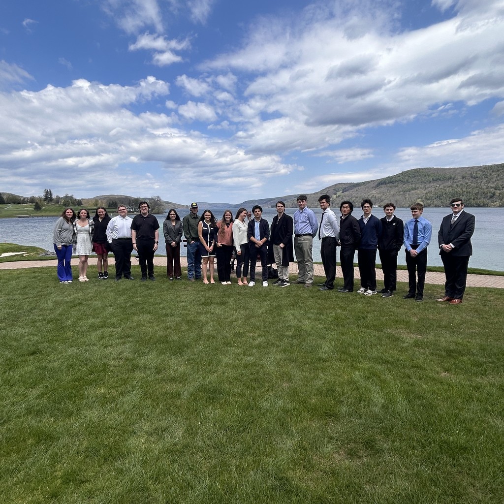 19 students standing on grass in front of a lake with mountains behind them.
