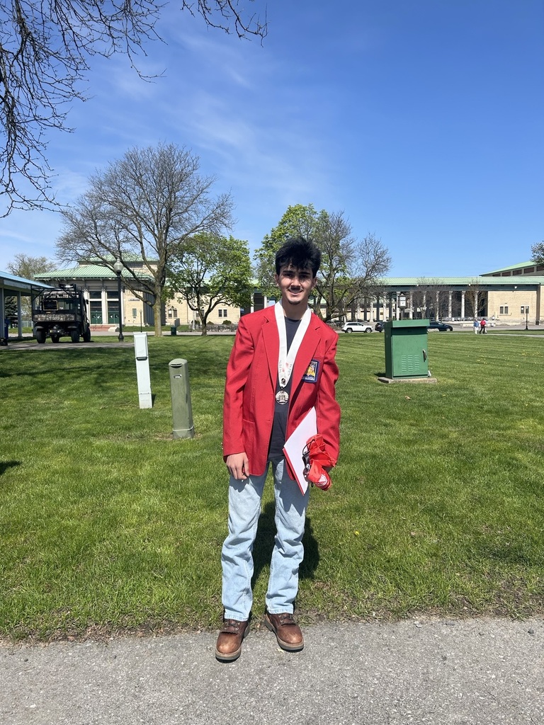 Student standing outside wearing jeans, work boots, black shirt and a red jacket. He is also wearing a medal.