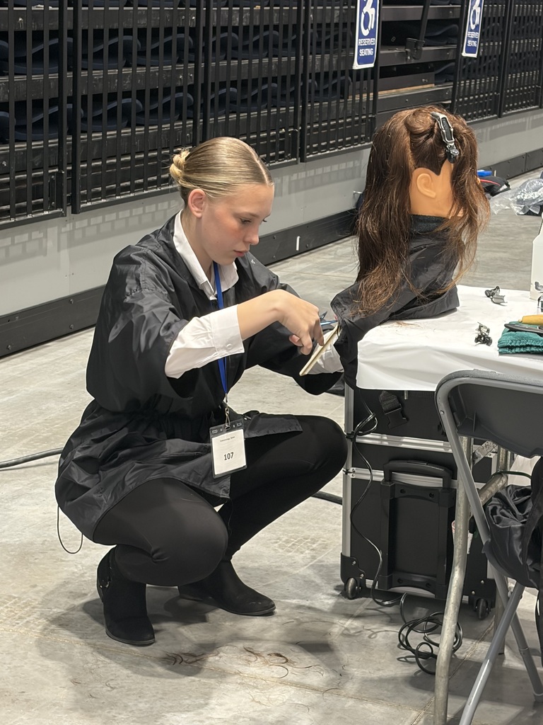 Student wearing black pants, white shirt, and black smock crouching down doing a hair cut on a mannequin head