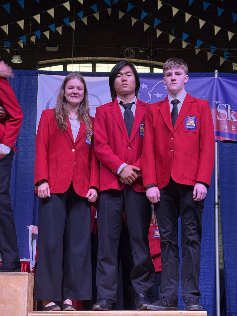 Three students standing on a stage wearing red jackets and black pants.