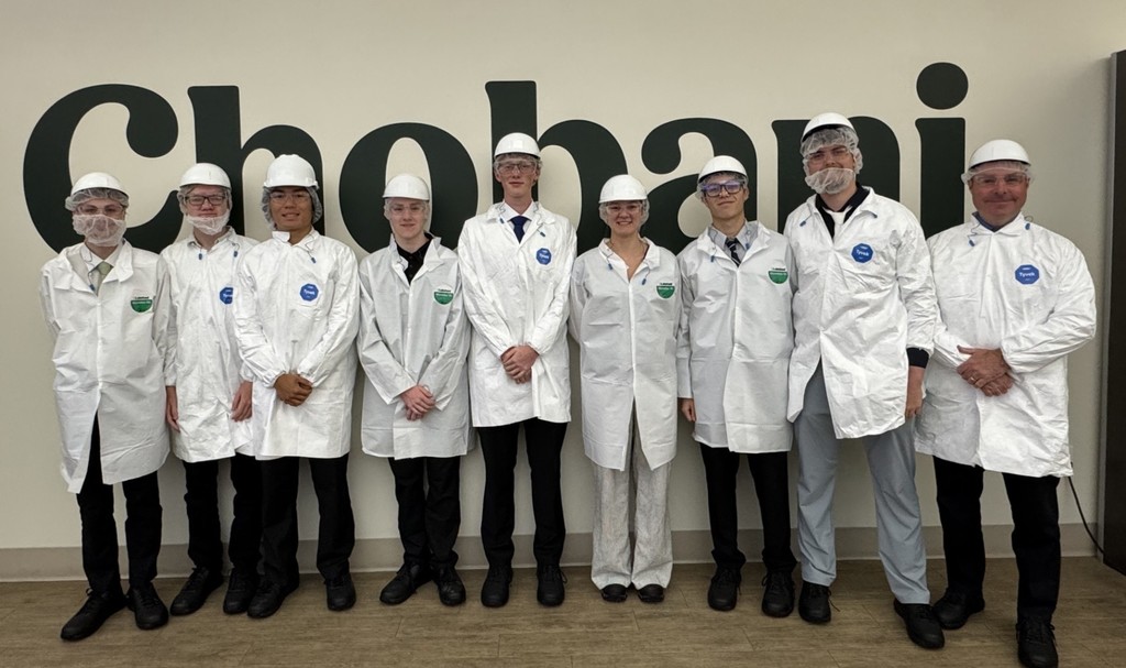 8 students and the NV Engineering instructor, in white jackets, white hard hats, and face protection standing in front of a Chobani sign at the plant.