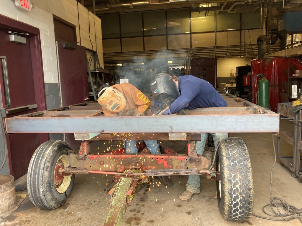 Two students wearing welding helmets welding pieces of a tractor.