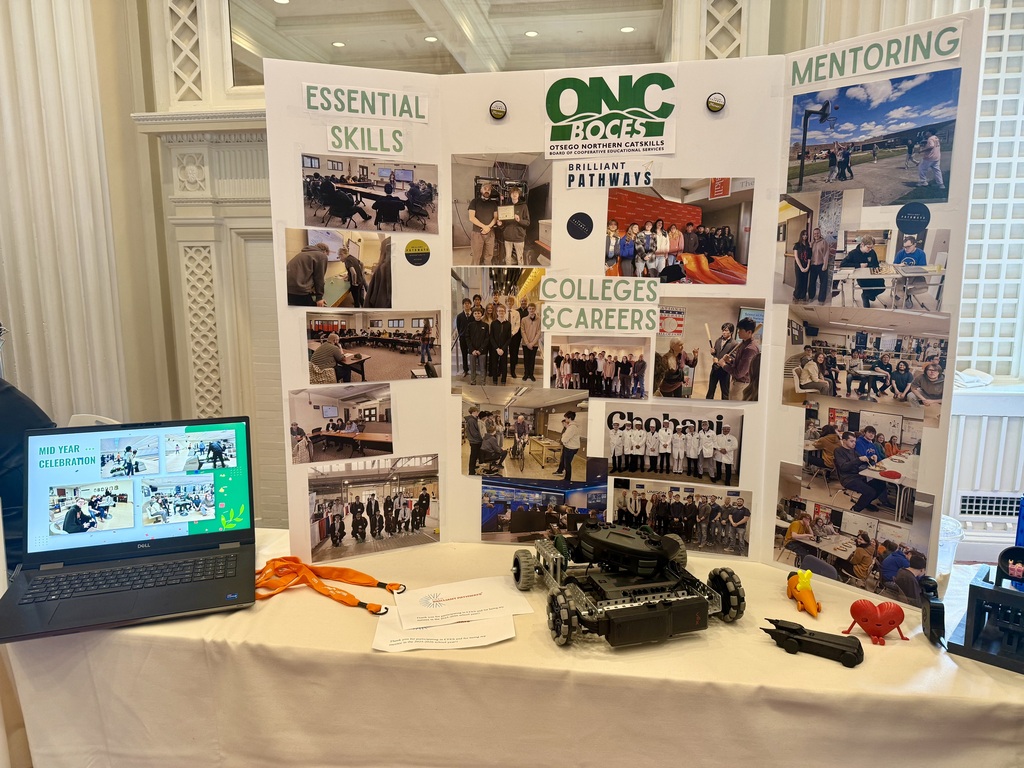 Table with New Visions Engineering presentation supplies, including a poster board that says Essential Skills, College and Careers, Mentoring with photos of the class doing different things. There's also a laptop other small items on the table.