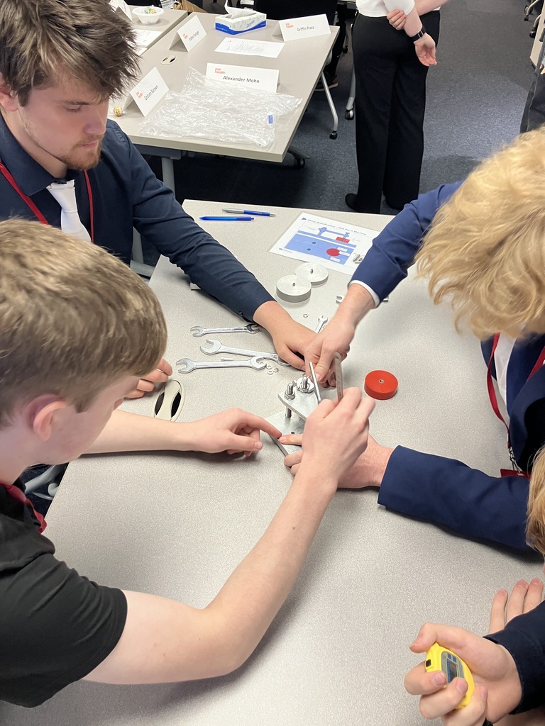 Four students sitting at a table, one with a stopwatch and the others working on solving something with tools.
