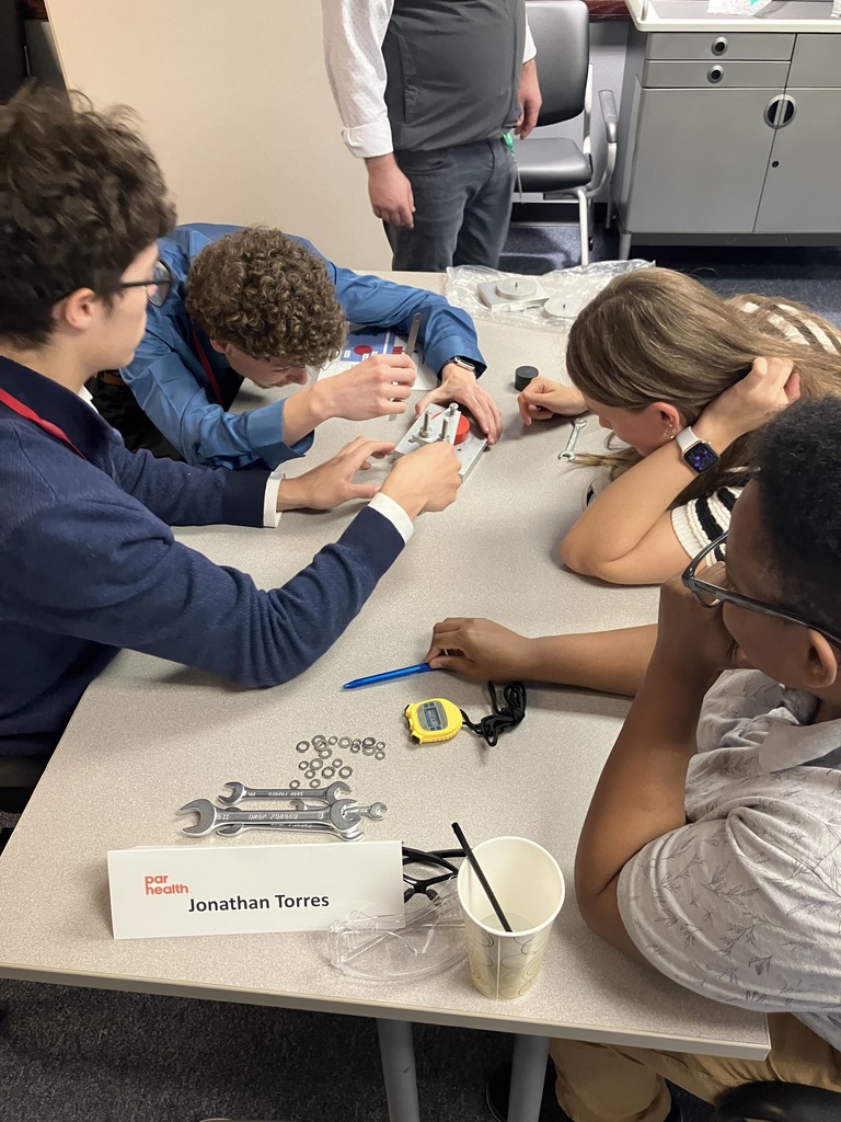 Four students sitting at a table, one with a stopwatch and the others working on solving something with tools.