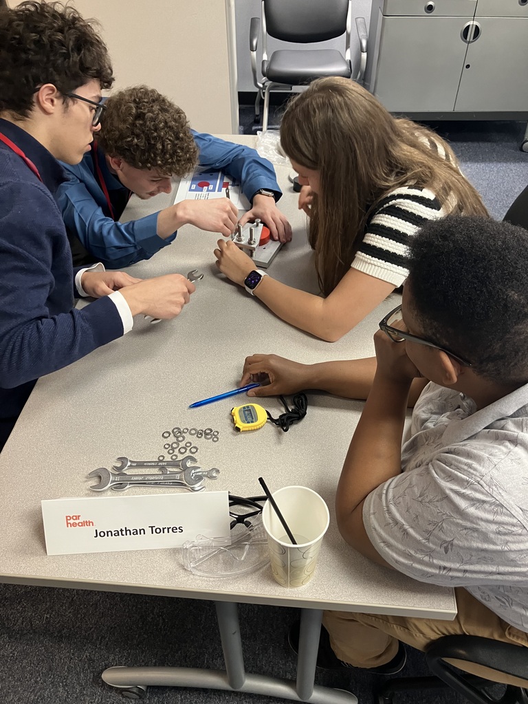 Four students sitting at a table, one with a stopwatch and the others working on solving something with tools.