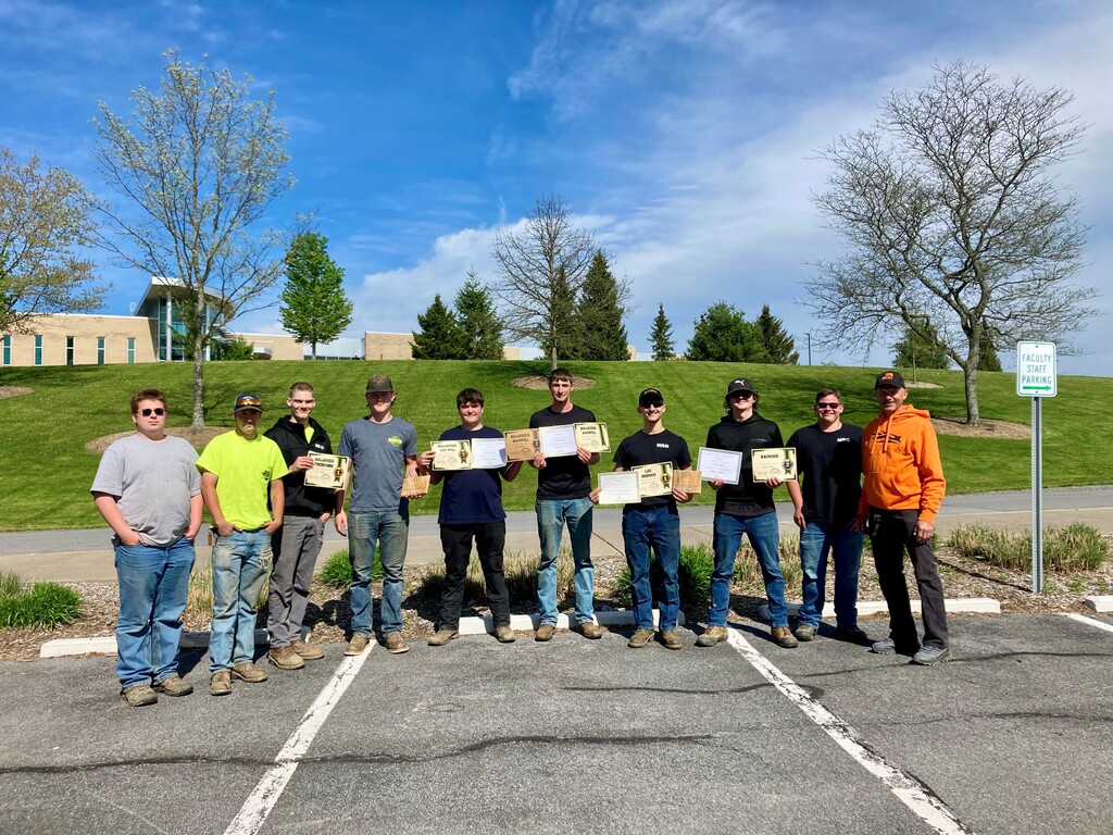 10 students standing in a parking lot. Some holding plaques and certificates.