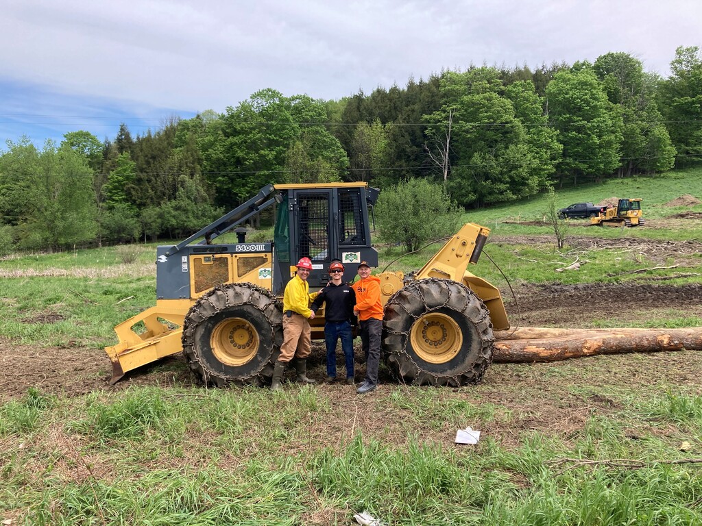 Students and adult standing in front of a bulldozer with logs in the background