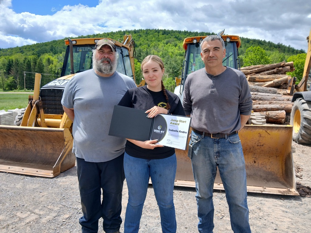 Student and two instructors standing in front of two bulldozers. The student is holding her award.