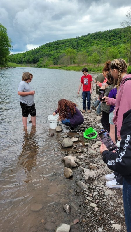 Students standing at the edge of the river. One student in the river with a cup and the teacher with a bucket.