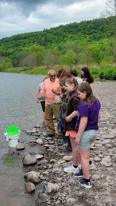 Students and adults standing on the edge of the river with a bucket.