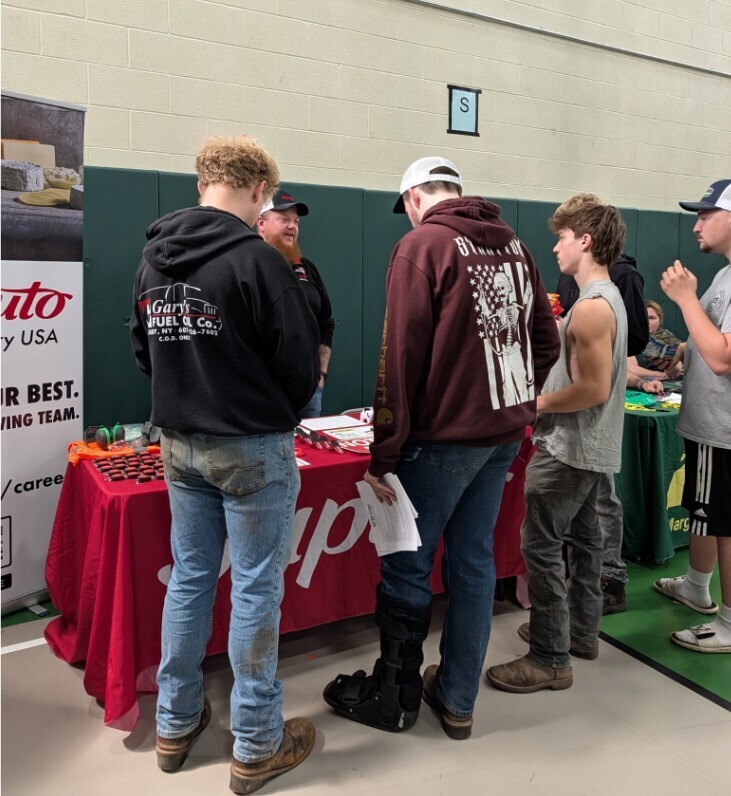 Four students speaking with a reprentative from Saputo. The table has a red tablecloth and white Saputa on it.