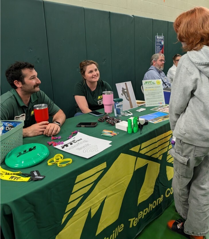 A student talking with two people sitting behind a table with a green tablecloth with yellow MTC Margaretville Telephone Company logo.