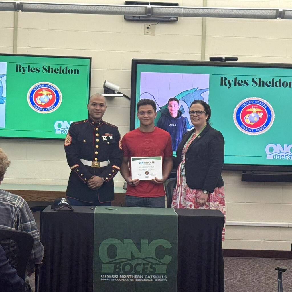 Marine recruiter, student holding a certificate, and ONC BOCES district superintendent posing behind a table with a black tablecloth and green ONC BOCES banner