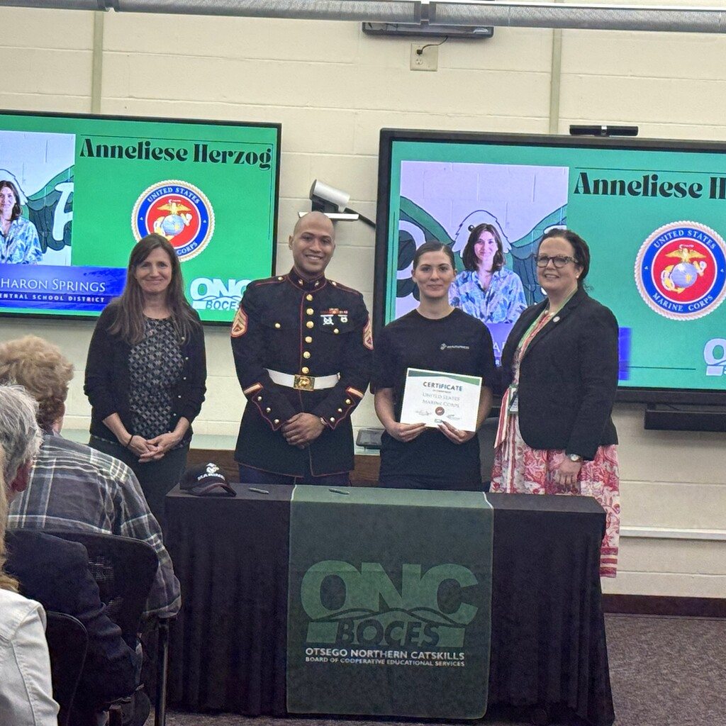 Marine recruiter, student holding a certificate, school superintendent, and ONC BOCES district superintendent standing behind a table with a black table cloth and green ONC BOCES banner. The student is holding a certificate.