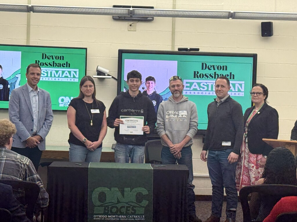 School superintendent, student's parents, student holding a certificate, and business representative posing for a picture behind a table with a black table cloth and green ONC BOCES banner.