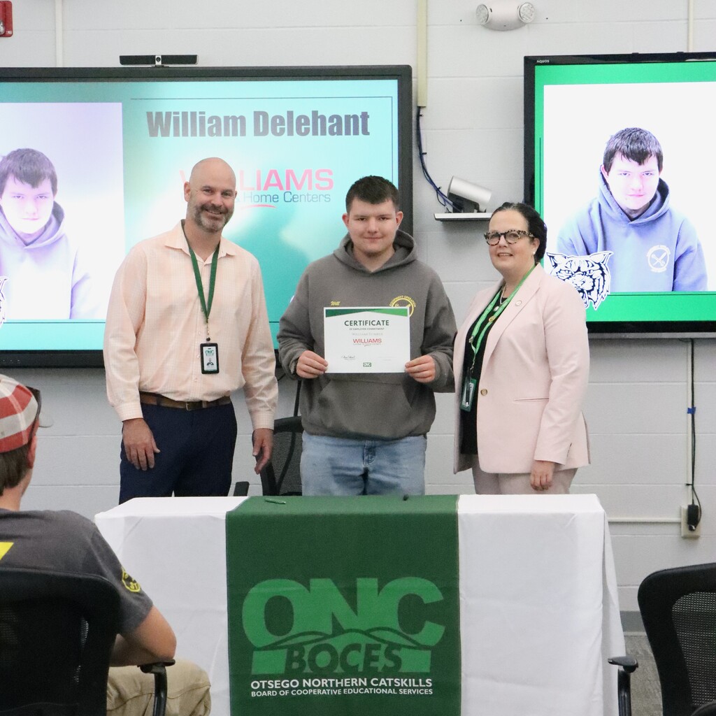 Assistant Superintendent, student, ONC BOCES district superintendent, standing behind a table with a white table cloth and green ONC BOCES banner. The student is holding a certificate.