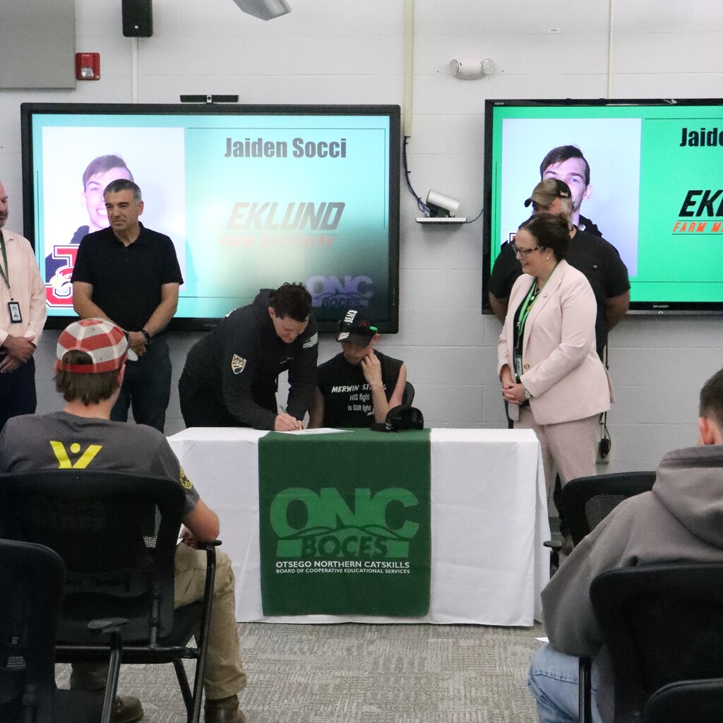 Assistant superintendent, instructor, ONC BOCES district superintendent watch as the employer signs the commitment letter next to the student who is sitting at a table with a white tablecloth with a green ONC BOCES banner.