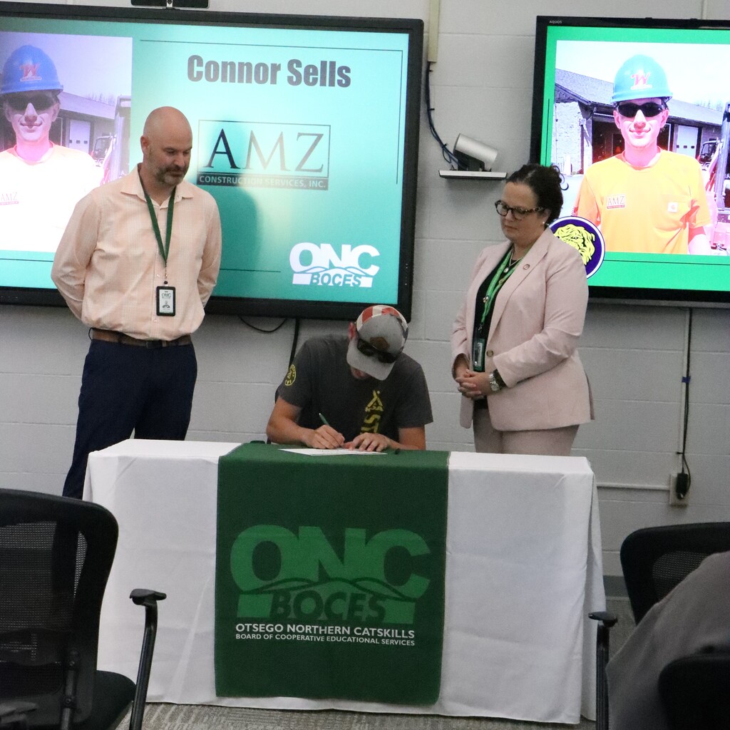 Assistant Superintendent and ONC BOCES district superintendent are standing as the student is signing the commitment letter on a table with a white tablecloth and green ONC BOCES banner.