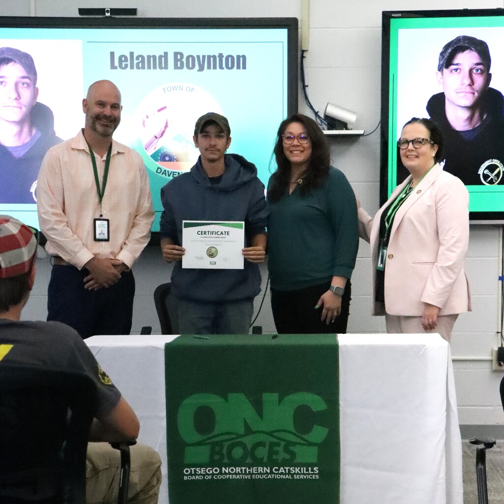 Assistant Superintendent, student, ONC BOCES district superintendent, and school superintendent standing behind a table with a white table cloth and green ONC BOCES banner. The student is holding a certificate.