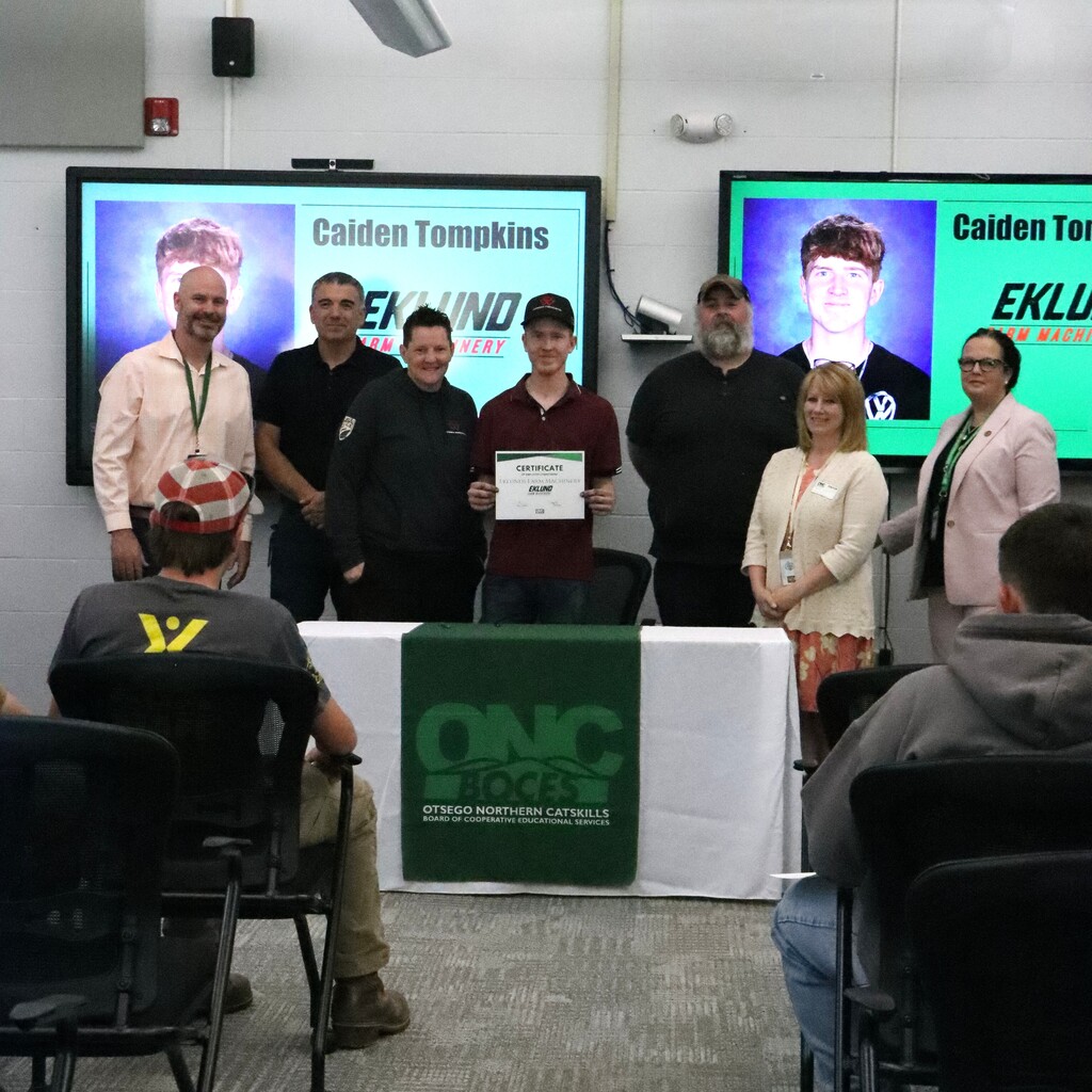 Assistant Superintendent, student, ONC BOCES district superintendent, two instructors, employer representative, and home school representative are standing behind a table with a white table cloth and green ONC BOCES banner. The student is holding a certificate.