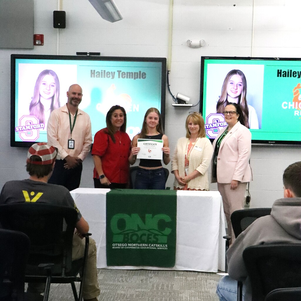 Assistant Superintendent, student, ONC BOCES district superintendent, instructor, and home school representative are standing behind a table with a white table cloth and green ONC BOCES banner. The student is holding a certificate.