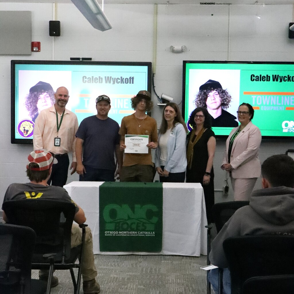 Assistant Superintendent, student, ONC BOCES district superintendent, instructor, parent and home school representative standing behind a table with a white table cloth and green ONC BOCES banner. The student is holding a certificate.