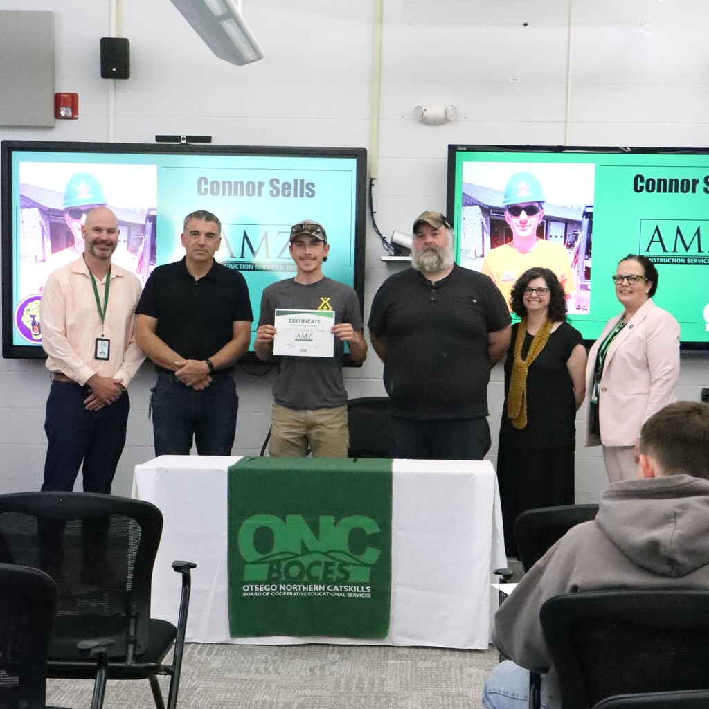 Assistant Superintendent, student, two instructors, ONC BOCES district superintendent, and school home school representative are standing behind a table with a white table cloth and green ONC BOCES banner. The student is holding a certificate.