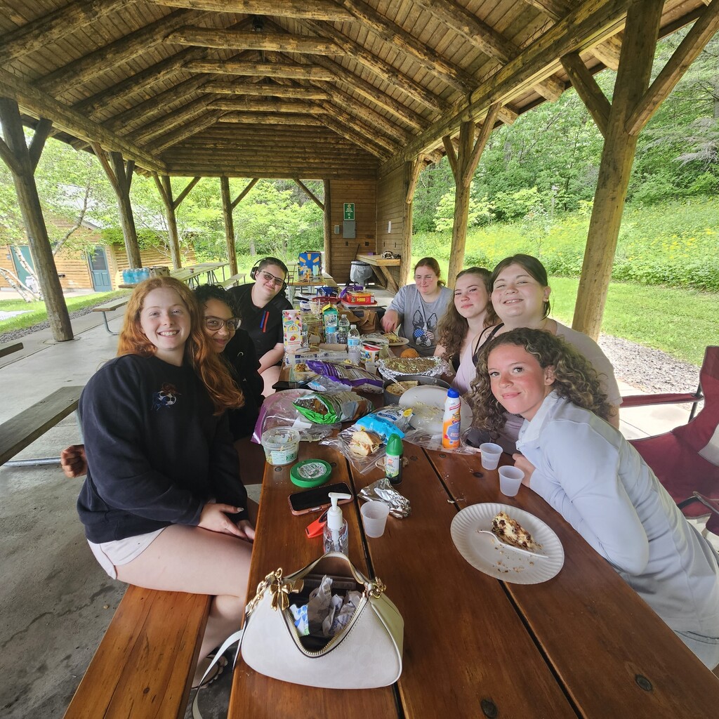 7 students sitting at a picnic tables with food on them. Students are posing and smiling for the picture.