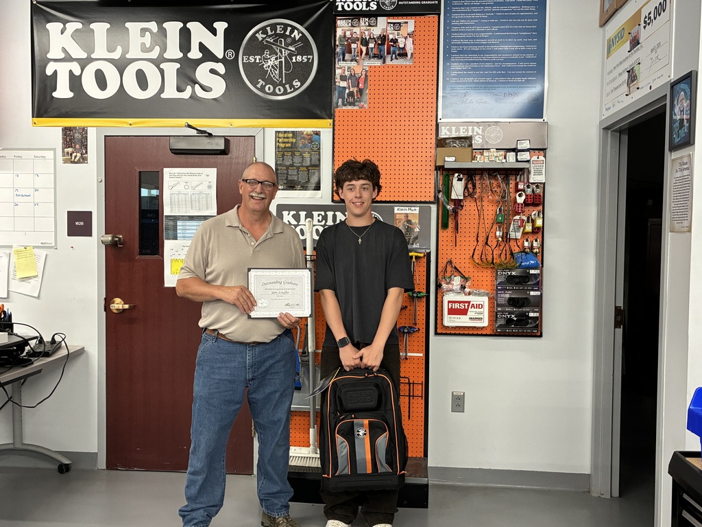 Student and instructor posing for a picture in the Mechatonics lab with a Klein Tools banner on the wall behind them. The student is holding the backpack of tools and the instructor is holding a certificate.