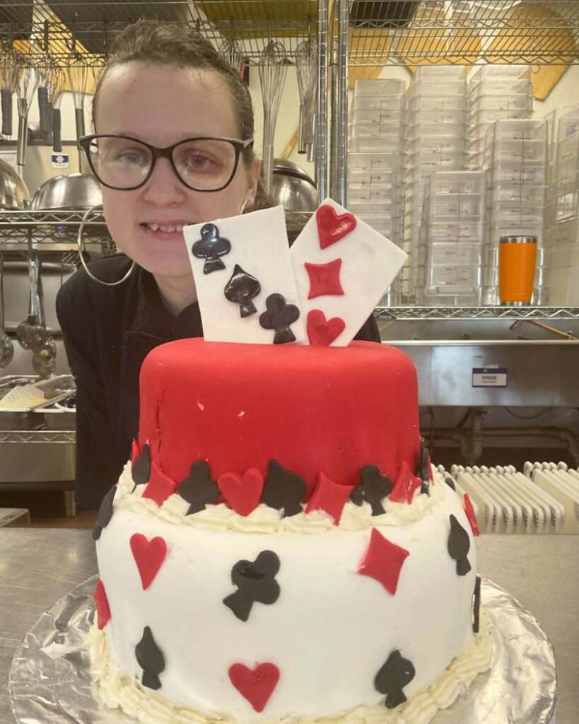Student standing behind a two tiered cake with one tier being red and one white. It has the playing cards symbols on it and two fondant playing cards on the top.