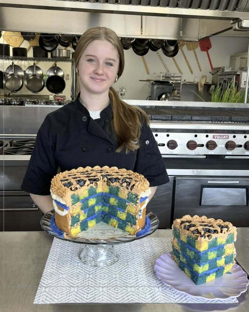 Student standing behind their cake with a large slice cut to show the checkerboard inside. The cake is decorated like a blueberry pie.