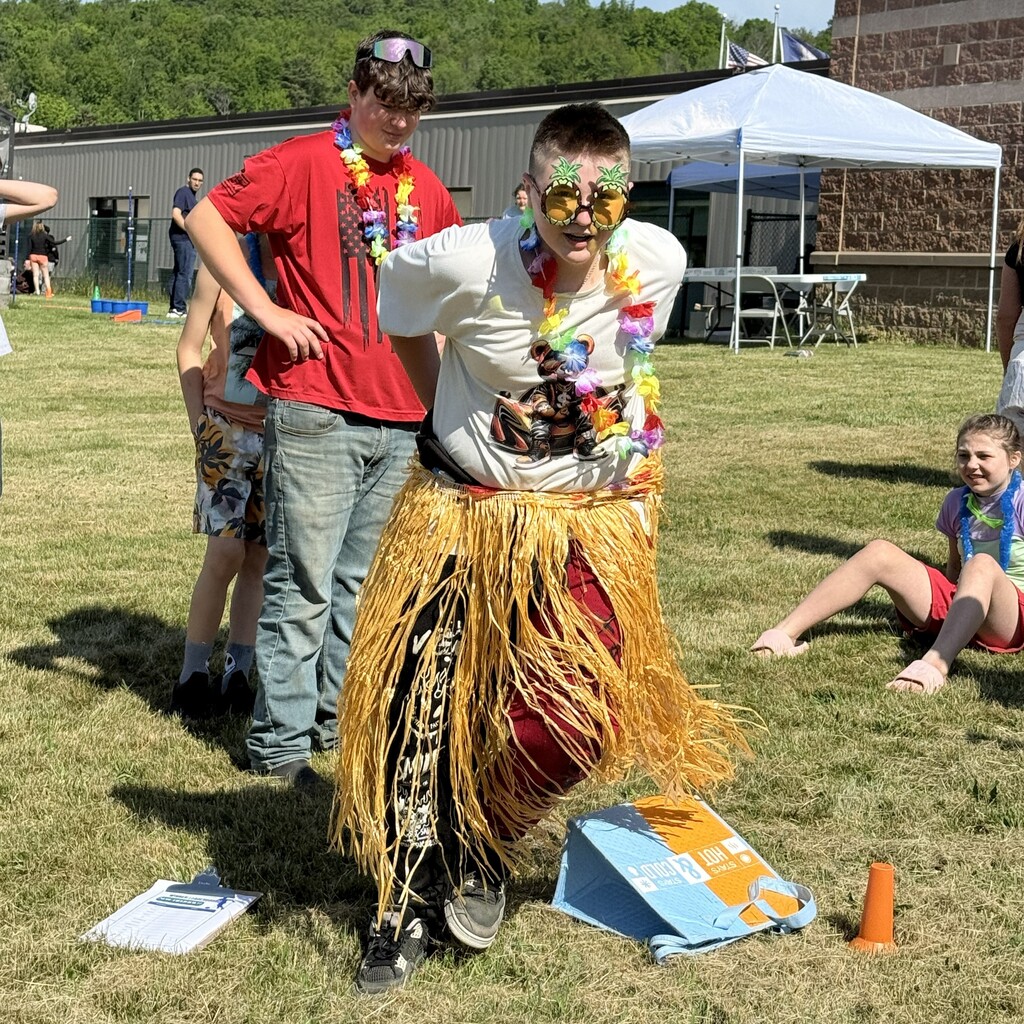 One student wearing a grass skirt, plastic lei, and sunglasses starting to run while other students look on