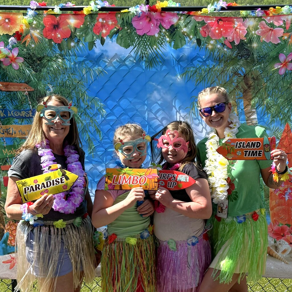 Two staff members and two elementary students posing in front of a Hawaiian themed photo backdrop. Each person is wearing grass skirts and holding signs that say, Paradise, Limbo, Surf's up, and island time.