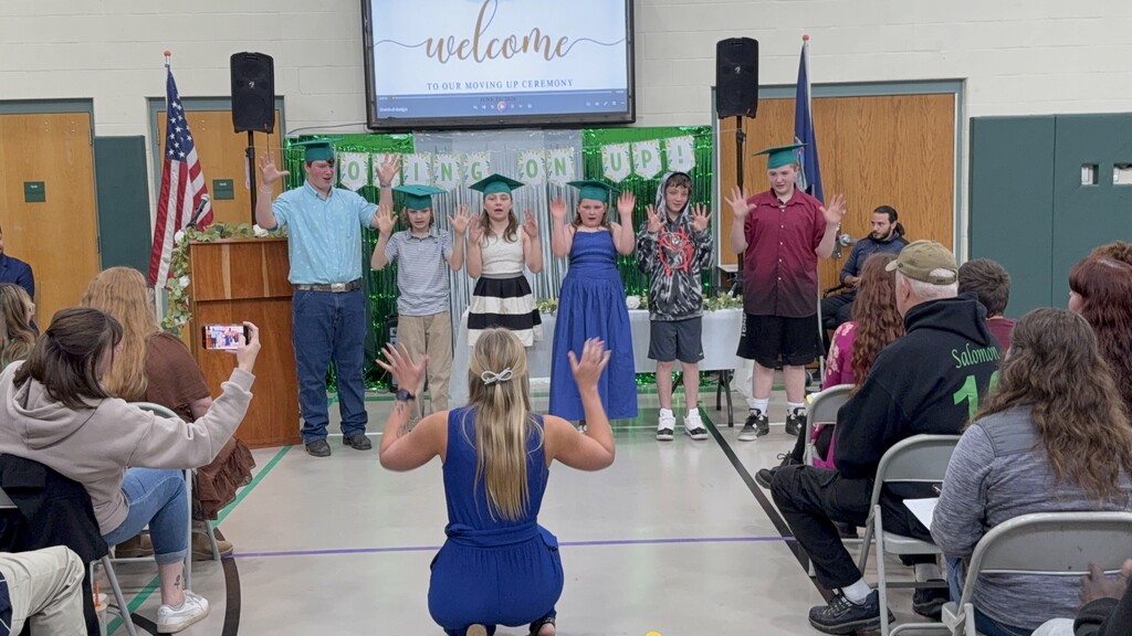 6 middle school students standing in the gym in front of guests performing a song with green graduation caps and tassles