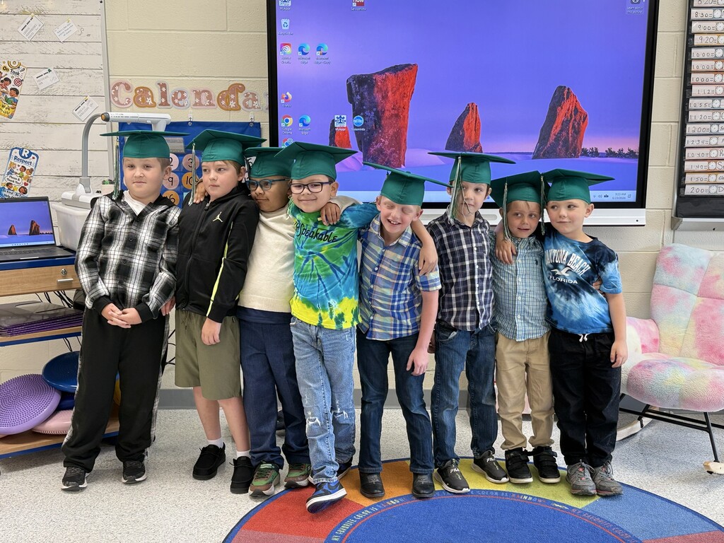 8 elementary students standing together with arms around each other with green graduation caps and tassles