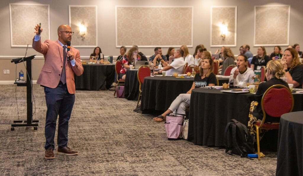 Person holding a microphone speaking to a large group of people sitting in a room around smaller tables.