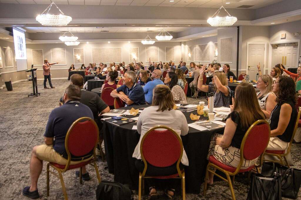 Picture of the large group of participants listening to the keynote speaker. They are seated at smaller tables in a large room.