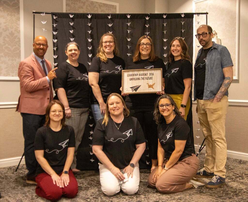 8 people posing with the keynote speaker with one person holding a sign that says Leadership Academy 2026 Unfolding the future, with a picture of a paper airplane and a origami crane