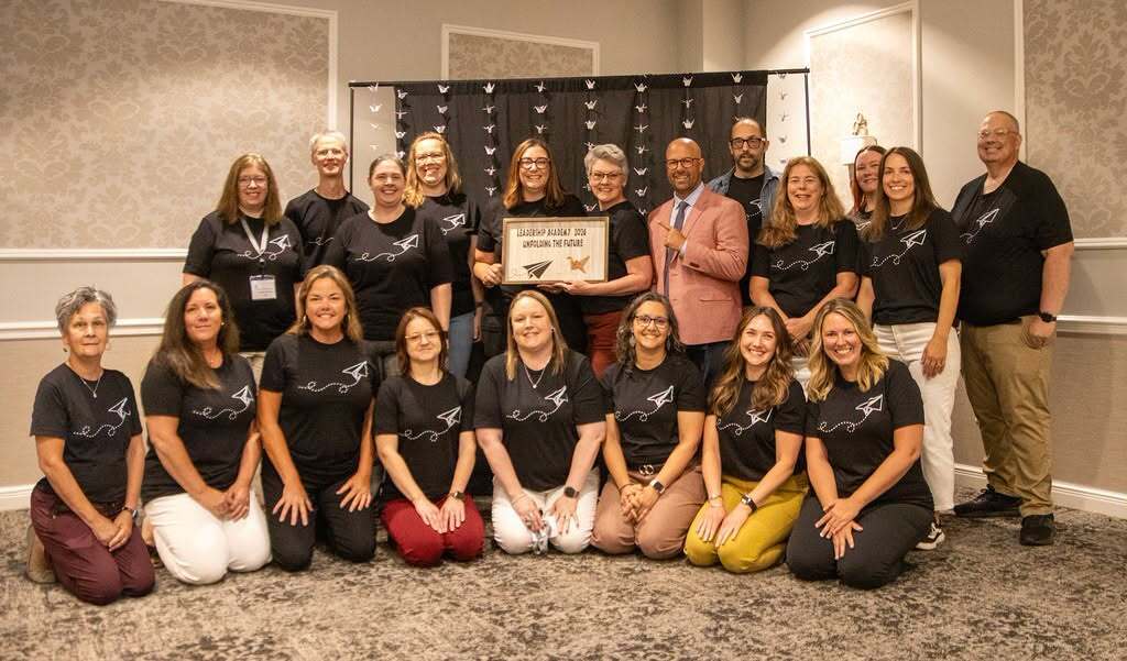 19 people posing for a picture with the keynote speaker with one person holding a sign that says Leadership Academy 2026 Unfolding the future, with a picture of a paper airplane and a origami crane