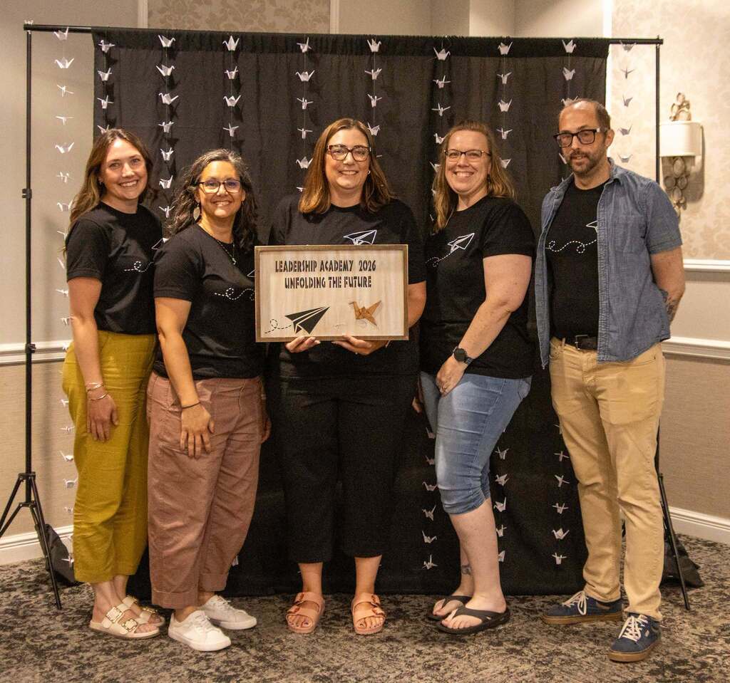 Group of 5 people posing for a picture with one person holding a sign that says Leadership Academy 2026 Unfolding the future, with a picture of a paper airplane and a origami crane