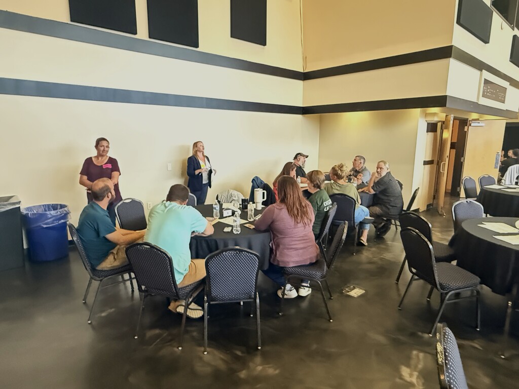 Two people standing in front of a room with 9 people at tables listening.