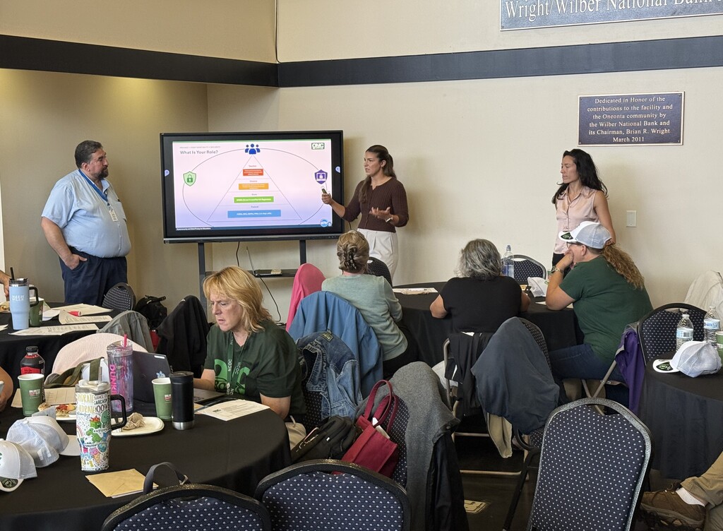 Three presenters standing with a small screen discussing cybersecurity with a small group of people sitting at tables.